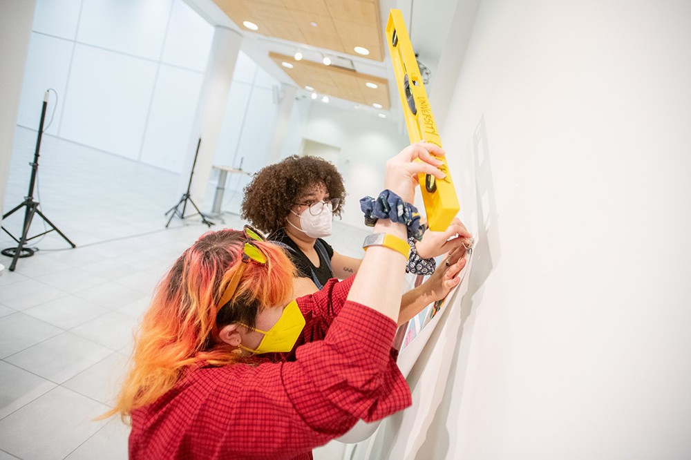 two students hanging artwork on a white wall.