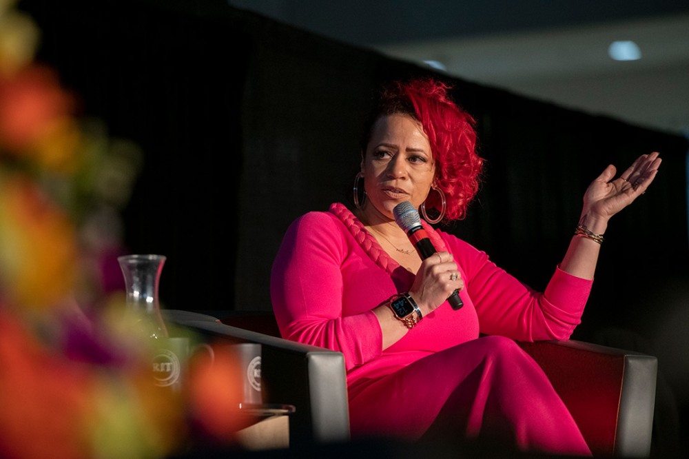 person sitting in an armchair on a stage speaking into a microphone.