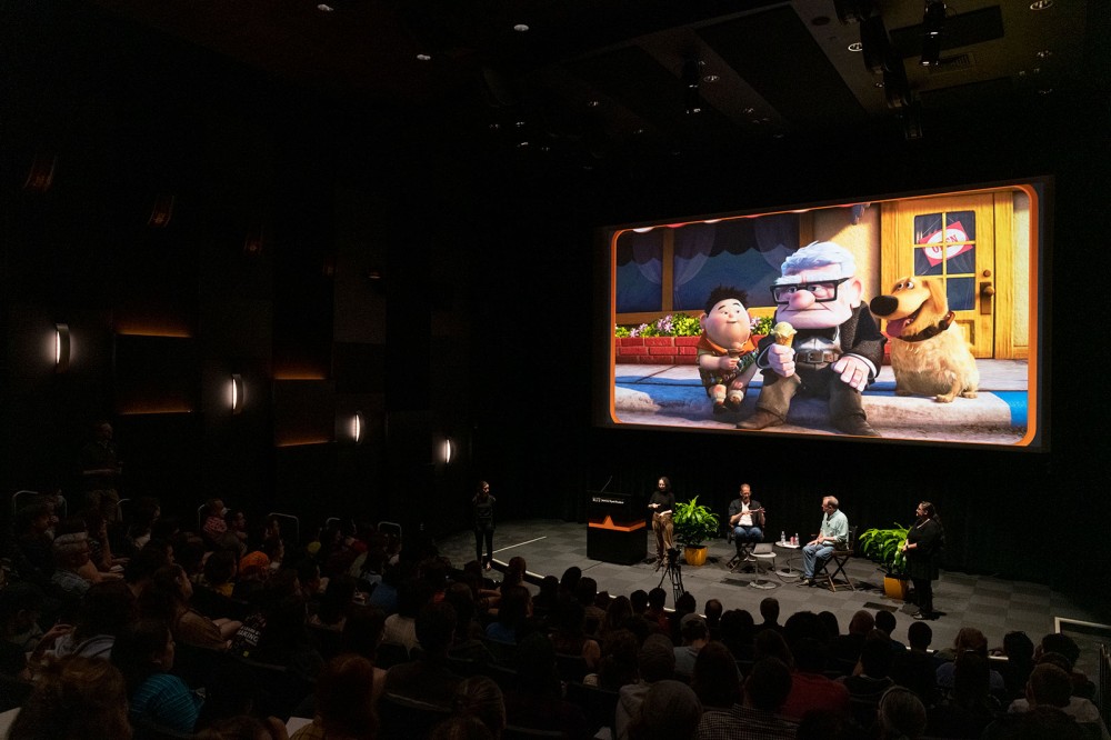 a dark auditorium with four people on stage and a scene from the movie Up projected above them.