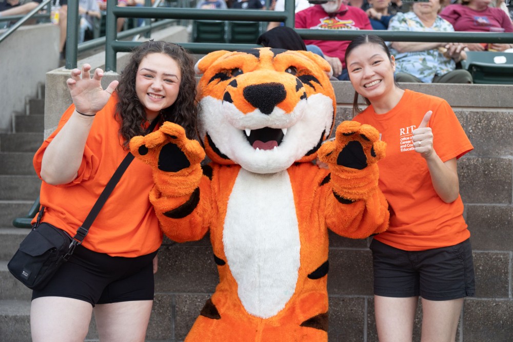 Two students dressed in orange pose with RIT mascot Ritchie.