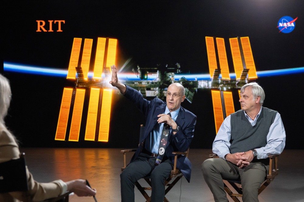 Man gestures while speaking during interview, seated beside colleague, with ISS solar arrays and NASA and RIT logos in background