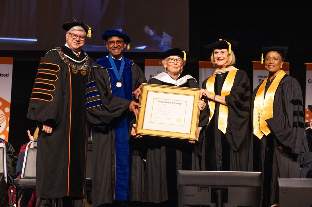 A group of people in academic regalia stand on stage, with one holding an honorary diploma