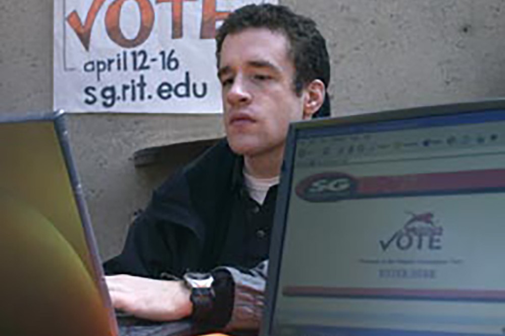 A student sits in front of a laptop with a Vote sign behind him.