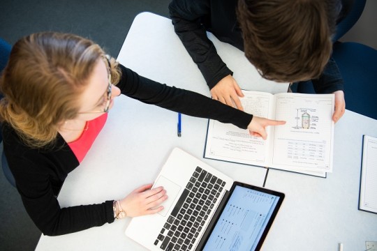 Overhead shot of two students. One student is seated at a computer pointing to a visual in the notebook of the other student.
