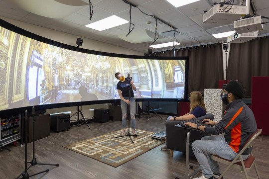 Student stands in front of super wide screen in lab displaying the inside of a mansion while other students observe.