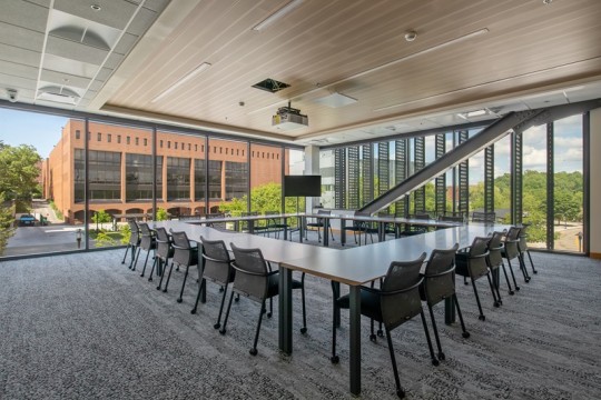 a large meeting space surrounded by floor to ceiling windows, and a large rectangular table with chairs in the middle.