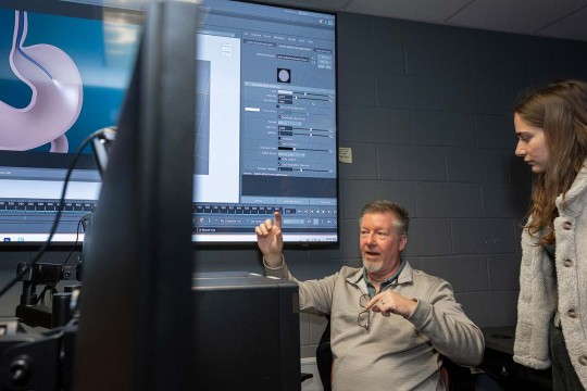 professor talks to a student in a lab with a large screen showing a digital image of a stomach.