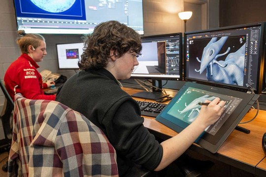a student draws on a tablet, which also displays on a computer screen.