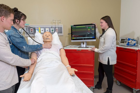 3 students using a stethoscope on a dummy patient and a monitor with data readouts