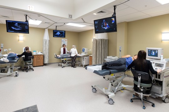 a hospital room with 3 patients all using ultrasound scanners with TVs on the wall showing the scan imagery