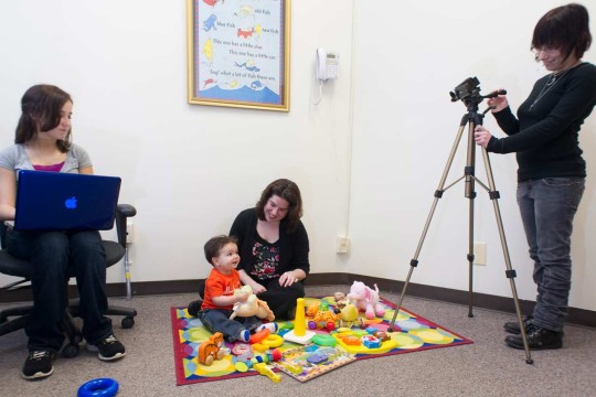 Two people recording a baby playing on a play mat with another adult