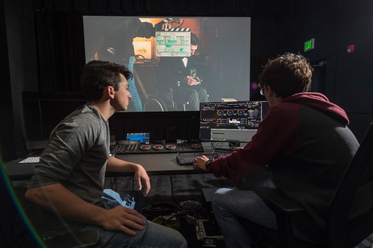 Two students sit in dark room in front of controls as screen plays movie in background