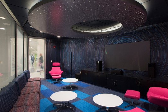 Dark room with white circular tables, hot pink chairs and large TV screen with speakers