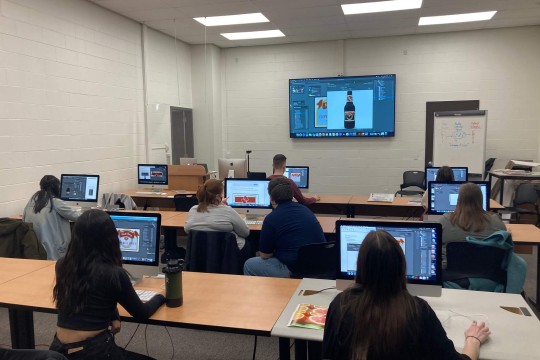 students in a computer lab look up at a T V displaying a label on a bottle.
