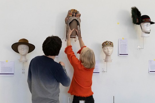 Two people placing headwear on display in a gallery