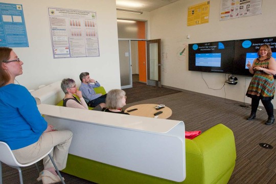 People on a couch watching a presentation on two televisions