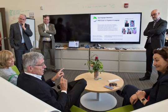 A group of people around 2 televisions talking while some sit and some stand