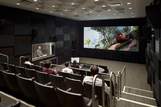 Student sits at table with controls while another student sits at electric keyboard and screen in background with controls projected onto it
