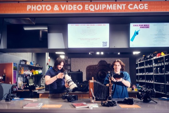 Two Cage employees behind the counter of the Cage, looking at camera equipment.