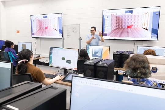 professor gesturing at front of computer lab, with two large T Vs displaying a chair in a room.