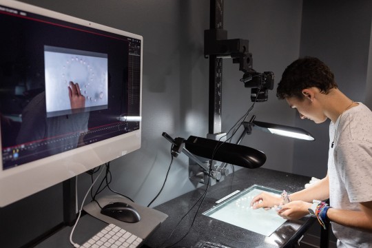 student places small white beads down on a glass surface with lights and a camera pointing at it. The image is displayed on a computer monitor.