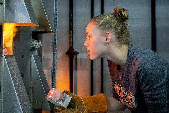 a student looks into a kiln while holding a ceramic brick with a protective leather glove.