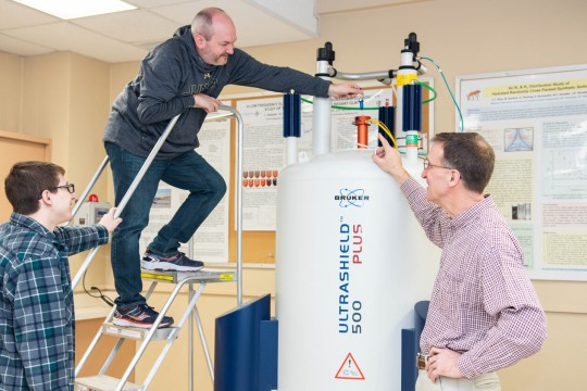 a man climbs a stepstool to reach the top of a Bruker Ultrashield 500 spectrometer, a large tank, as two others look on.