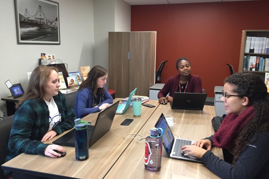 Three students and a faculty member working on laptops around a table.