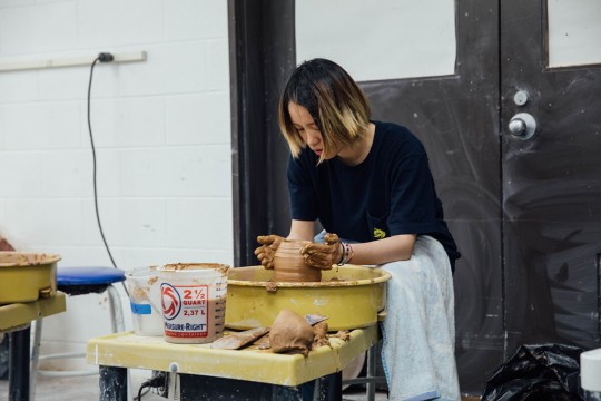 a student spins a tall clay vessel on a wheel.