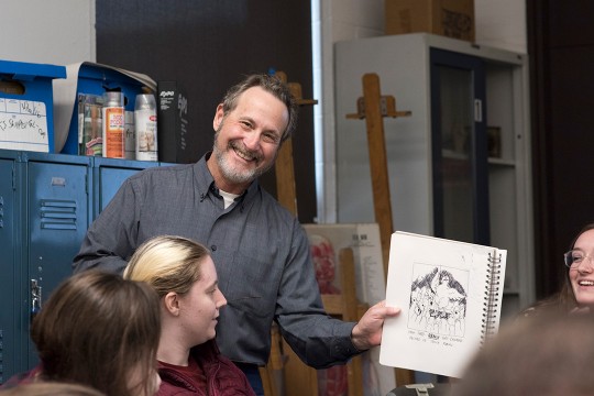 Leigh Rubin holds up a sketch and smiles at the camera as students look on.