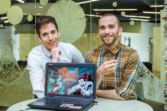 2 students sitting at a desk, showcasing their game on a laptop screen.