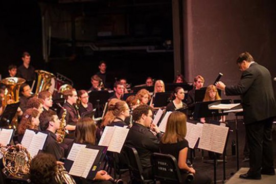 A photo of RIT's concert band performing. The conductor stands on a short platform at the front and uses his hands to instruct the players. 