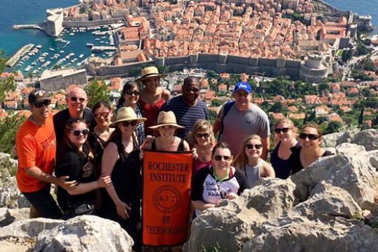 A group of RIT students and parents pose on the mountainside in Croatia, a beautiful city landscape behind them. They hold a banner that reads "Rochester Institute of Technology."