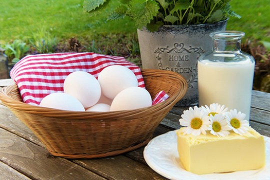 An artistic photo of eggs in a basket, milk in a glass bottle, and a large block of cheese with daisies on top of it.