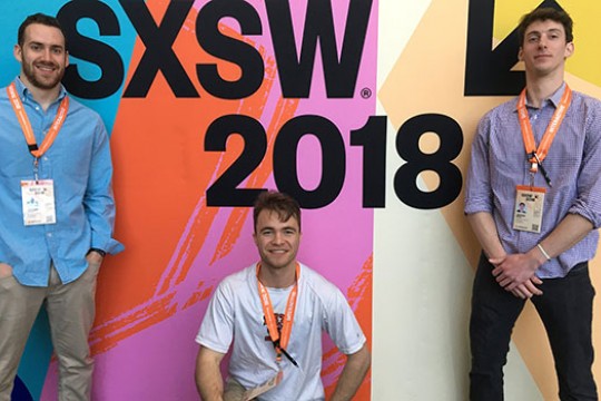 Three team members standing in front of a colorful wall that says "SXSW 2018".
