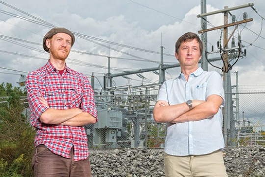 Eric Hittinger and Eric Williams post for a photo together in front of powerlines, both of their arms crossed.