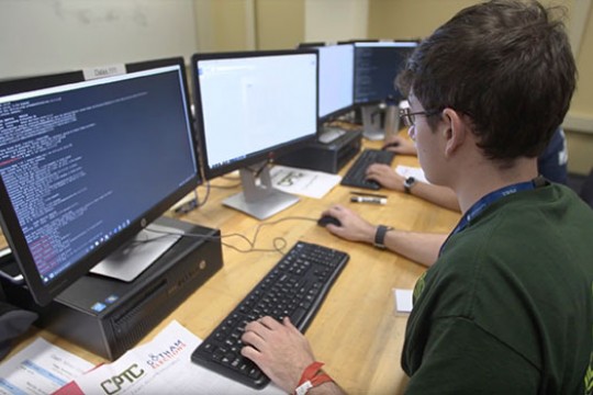 A student sits in front of a computer and writes out code, which is visible on the desktop screen.