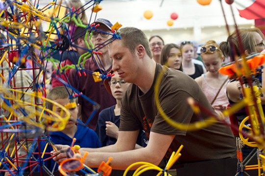 A student at Imagine RIT adjusts the toy-model of a theme park while children and their parents look on behind him.