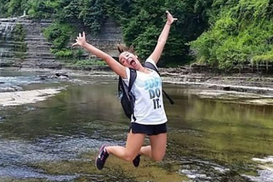 Nina Hullett jumps up in the air, extending her arms and smiling wide. She is outside near a large creek, seemingly on a hike.