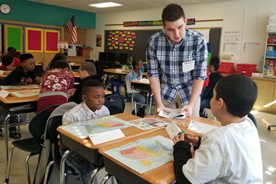 A person talking to two kids in a classroom. Kids are sitting at desks with maps of America in front of them.