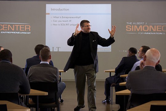 Richard DeMartino with hands up lecturing in front of people.