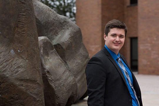 Zachary smiles at the camera, leaning against an outdoor sculpture behind the College of Liberal Arts.