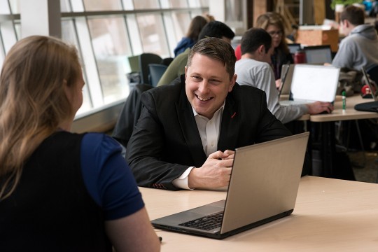 Michael Oshetski sits with a student in front of a laptop smiling.