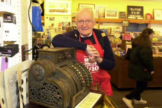 Older man wearing red apron poses with antique cash register