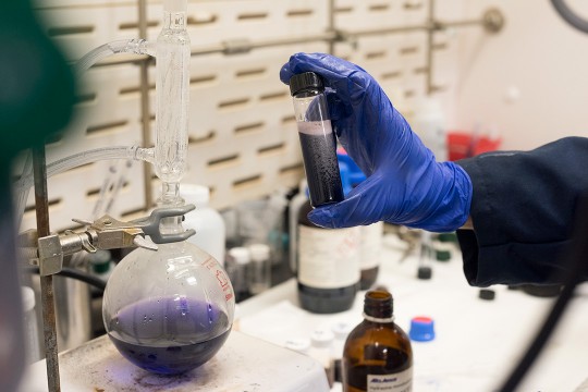 Gloved hand hold testtube of black liquid; container with purple liquid in background