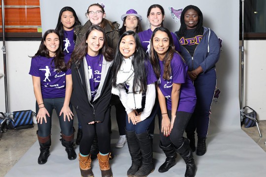 Group of nine female students wearing purple T-shirts stands together in front of white backdrop