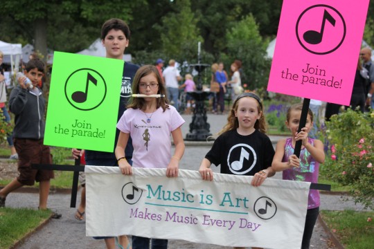 Community members hold a banner saying "Music is Art" with the subtext "Makes music every day". Students flanking the banner hold sings with the text "Join the parade"