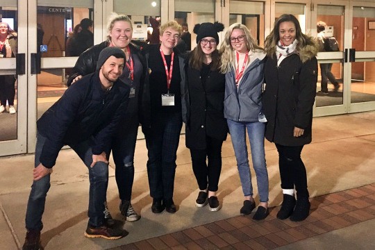 Group of six students stands outside of building with glass doors