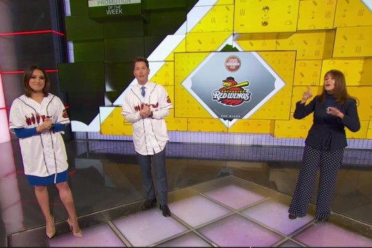 Two sports casters wearing Rochester Red Wings jerseys stand on set with interpreter.