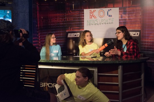 Three students sit at news desk, with one holding an inflatable microphone up to another.